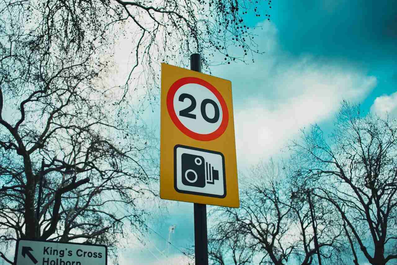 Speed limit sign showing 20 mph with camera warning in a leafy urban area.